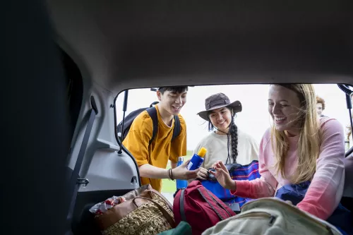 Friends wearing sun protection (long shirts, hats and sunscreen) are gathering their items from the back of their roadtrip car.